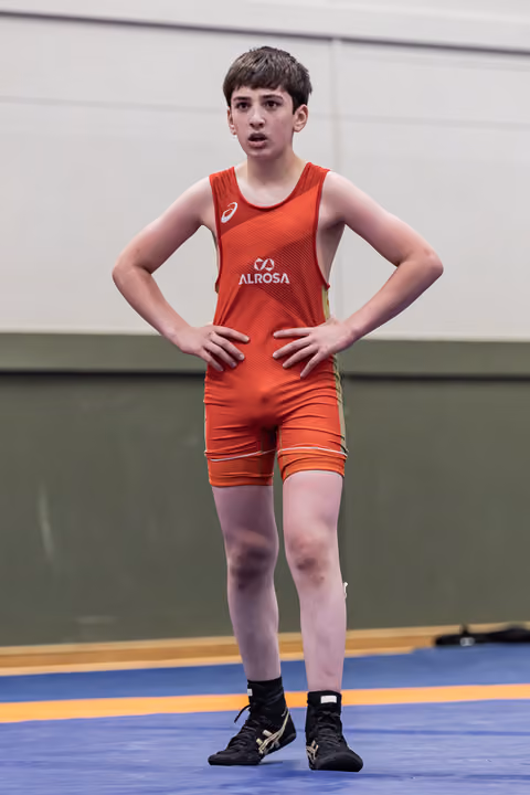 Young male wrestler in red Alrosa singlet stands hands on hips, alert expression, on blue competition mat.