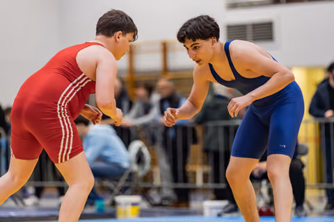 Two young wrestlers in red and blue singlets square off, crouched in tense ready positions on an indoor mat.