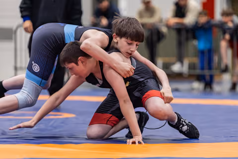 Two young boys wrestle intensely on a blue and orange mat, one in black dominating with focused determination.