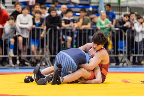 Wrestler in red singlet dominates opponent on yellow mat, crowd watching intently in background.