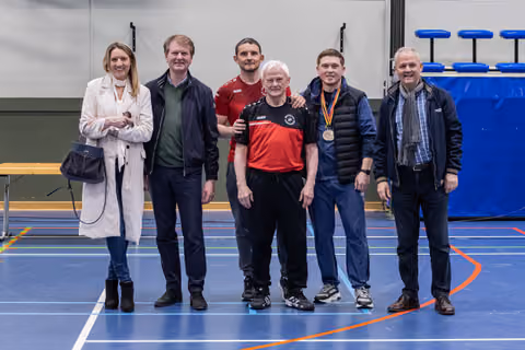 Six people pose proudly in a sports hall; a young man wears a bronze medal around his neck.