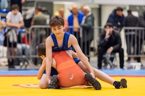Boy in blue singlet dominates opponent in red, pinning him to the yellow mat with focused concentration.