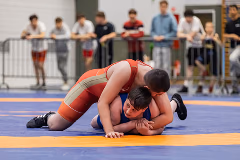 Young wrestler in red singlet pins opponent in blue to the mat while spectators watch from behind barriers.