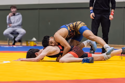 Blue-suited wrestler pins opponent face-down on yellow mat, referee watching nearby, spectator seated in background.
