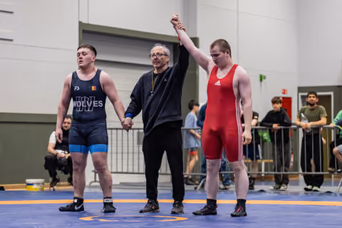 Referee raises the arm of a red-singlet wrestler declaring victory, while the defeated blue-singlet opponent stands subdued.