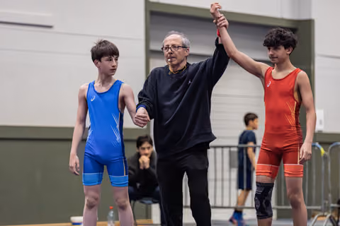 Referee raises the arm of the red-suited winner while the blue-suited boy stands stoic beside them in a gym.
