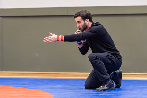 Male referee kneels on blue mat, blowing whistle and gesturing with outstretched arm during a wrestling match.