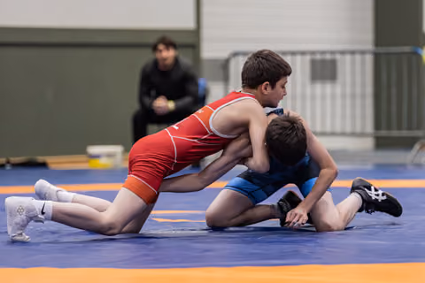 Two youth wrestlers compete intensely on a blue mat, one in red dominating from above as a coach watches from behind.