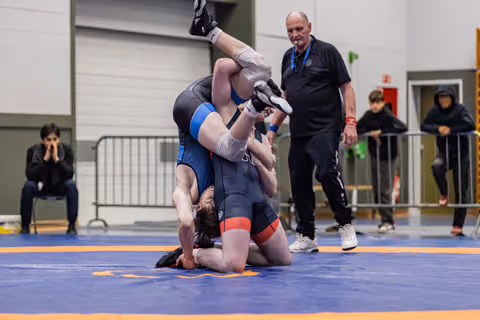 Two wrestlers locked in a hold on the mat while a bald referee watches closely, smiling, in an indoor sports hall.