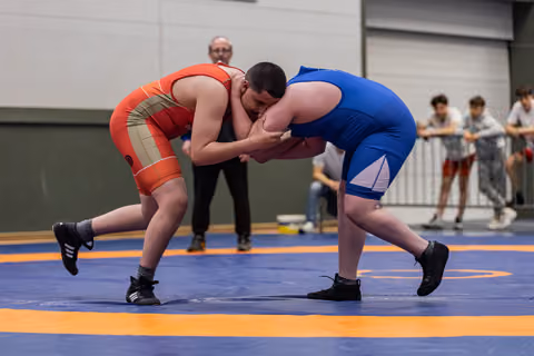 Two heavyweight wrestlers locked in a tight clinch on a blue mat, referee watching closely behind them.