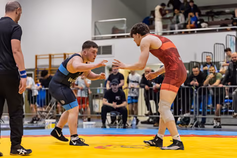 Two wrestlers in navy and red singlets face off in a tense stance, watched by a crowd at an indoor sports hall.