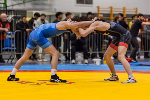 Two wrestlers lock heads in a tie-up stance on a yellow mat, crowds watching in the background.