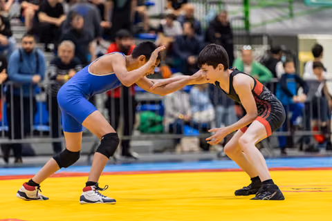Two young wrestlers in blue and black singlets square off in a tense stance, crowd watching in the background.
