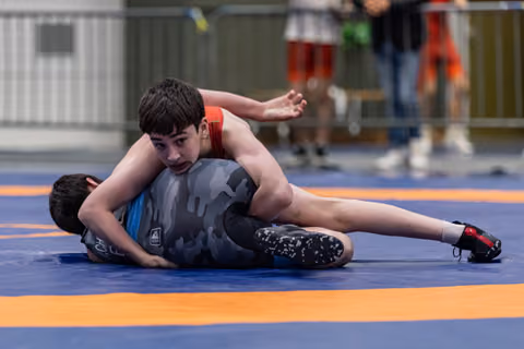 Teen wrestler in red singlet pins opponent on blue mat, focused expression, arm extended for balance.