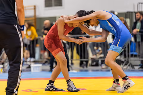 Two young female wrestlers in red and blue singlets lock hands in an intense stance on a yellow competition mat.