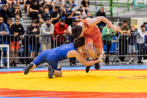 Blue wrestler launches red opponent airborne in dynamic throw, crowd watching intently in packed arena
