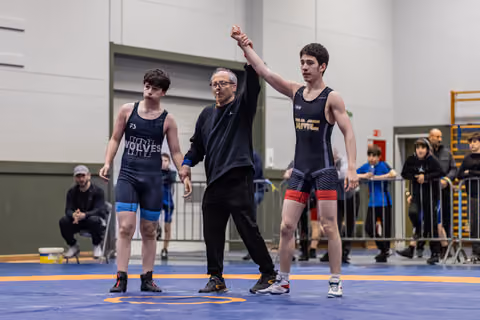 Referee raises the arm of the victorious wrestler in a black singlet, while the defeated opponent stands beside them.
