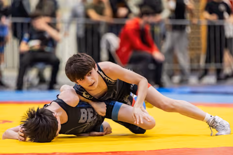 Focused teen wrestler dominates opponent on yellow mat, pressing down with determined expression during bout.