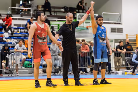 Referee raises blue-singlet wrestler's arm in victory while red-singlet opponent stands dejected on yellow mat.
