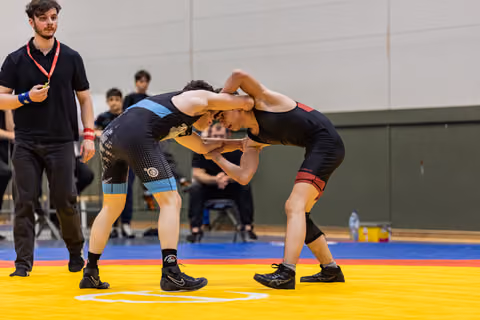 Two young wrestlers grapple head-to-head on a yellow mat, locked in a tense clinch, referee watching closely nearby.