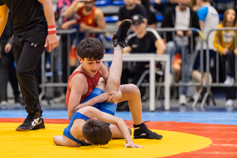 Young wrestler in red singlet grips opponent's leg on yellow mat, focused expression, referee watching nearby.