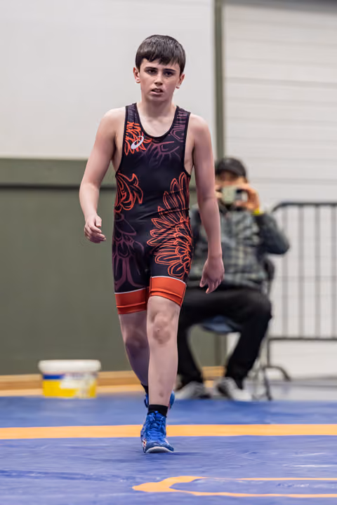Young boy in black and red singlet walks across the mat with a focused, determined expression during a wrestling event.