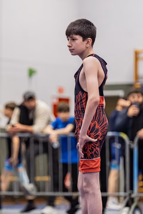 Young boy in red and black singlet stands alert and focused, waiting at the edge of the mat during a bout.