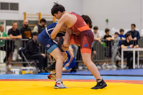 Two young wrestlers in blue and red singlets locked in a powerful clinch on a yellow mat, crowd watching behind.