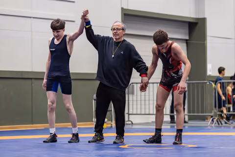 Referee raises the hand of a young wrestler in blue, declaring him winner, as his opponent in red bows in defeat.