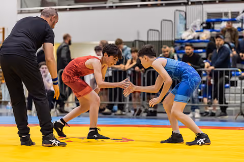 Two young wrestlers crouch in fierce concentration, reaching toward each other on a yellow mat as a referee watches closely.