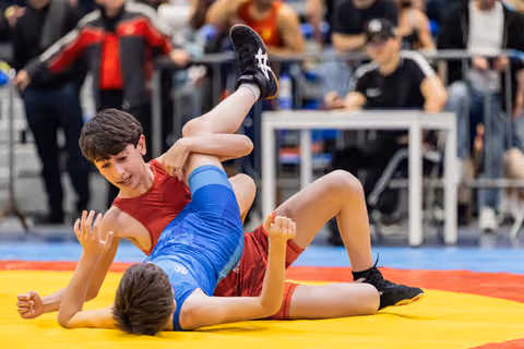 Young wrestler in red singlet dominates opponent in blue, lifting his leg on a colorful competition mat.