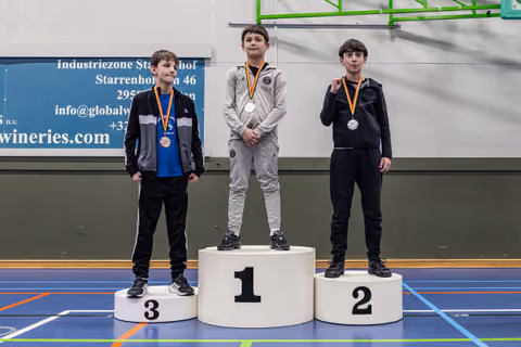 Three young boys stand on a podium, gold on top, silver and bronze beside, all wearing medals in an indoor gym.