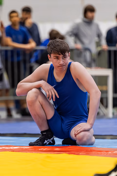 Teen wrestler in blue singlet crouches on the mat, looking intently at the camera with a calm, focused expression.