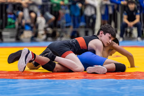 Young wrestler in black singlet controls opponent on the mat, focused expression during intense youth wrestling bout.