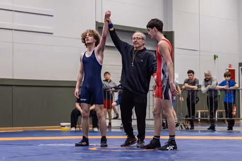 Referee raises the red-singlet wrestler's hand in victory while the blue-singlet opponent looks away, dejected.