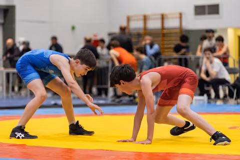 Two young male wrestlers in blue and red singlets crouch in tense readiness, eyes locked, on a yellow and red mat.