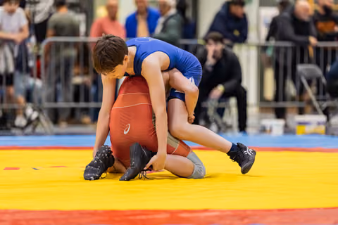 Young wrestler in blue singlet dominates opponent in red, gripping legs on a yellow mat with crowd watching.