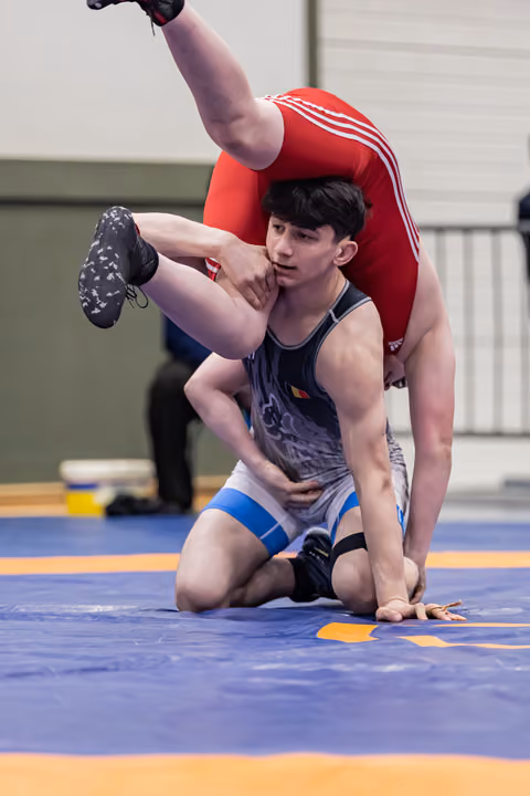 Focused teen wrestler lifts opponent overhead during match on blue mat inside sports hall.
