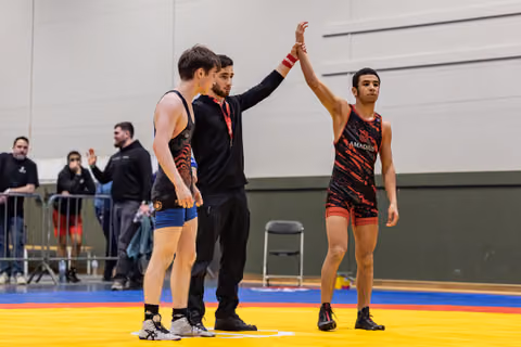 Referee raises the arm of an Amadius wrestler declaring victory, while the defeated opponent stands nearby on the mat.