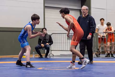 Two young wrestlers in blue and red singlets square off in fighting stance as a referee watches closely indoors.