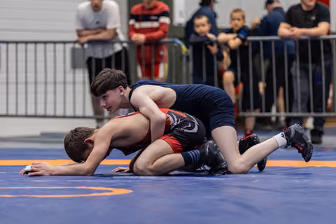 Young wrestler in navy singlet controls opponent on the mat, focused expression, spectators watching in background.