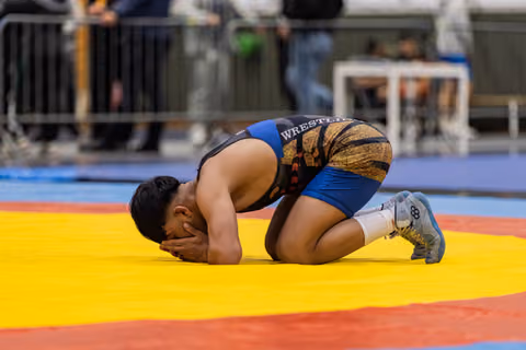 Young wrestler kneels on the mat, face buried in hands, expressing deep emotion after a match.
