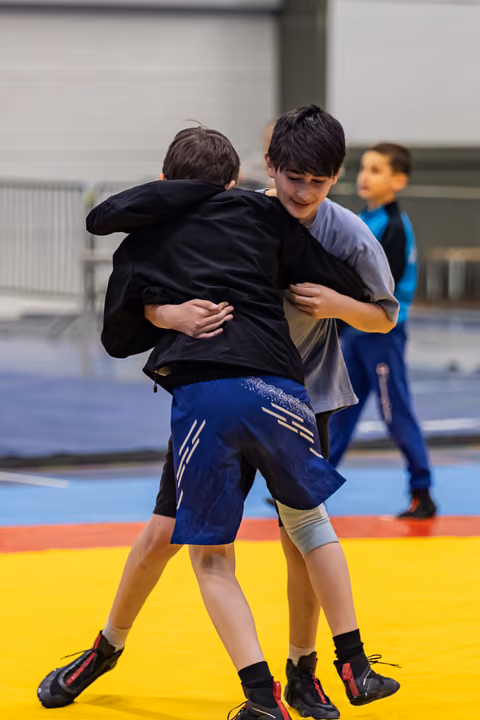 Two boys lock arms in a wrestling clinch on a yellow mat, one smiling, a third boy watches in the background.