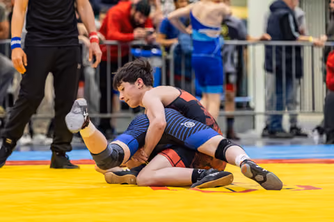 Young wrestler in red singlet controls opponent on yellow mat, pinning leg with focused determination at indoor tournament.