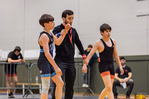 A coach in black gives focused instruction to two young wrestlers in a gymnasium, one in blue, one in black singlet.