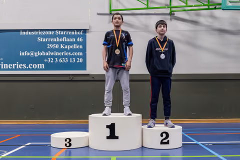 Two young boys stand on a podium, gold and silver medalists, looking serious in an indoor sports hall.