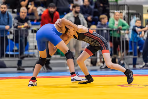 Two young wrestlers lock heads in an intense clinch, one in blue, one in black Bears singlet, on a yellow mat.