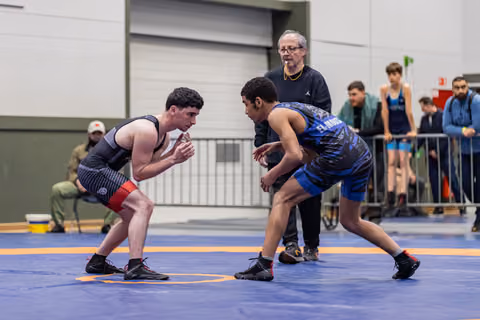 Two young wrestlers face off in intense stances on a blue mat, with a referee watching closely behind them.