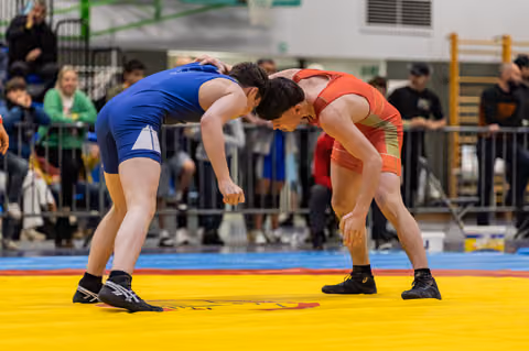 Two young wrestlers in blue and red singlets crouch in a tense standoff on a yellow competition mat, crowd watching.