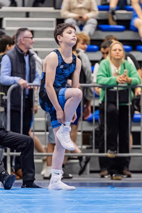 Young male wrestler stretches his leg with focused intensity, standing on a blue mat before his match.